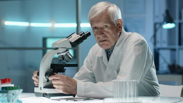 Senior male scientist working with microscope and then posing for camera at desk in laboratory