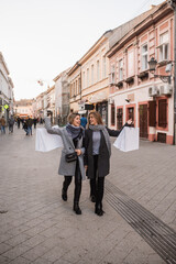Two beautiful fashion Caucasian women walk down the street smiling and carrying bags with purchased clothes. Autumn shopping in the city