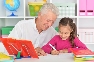 Cute girl doing homework with grandfather at the table at home