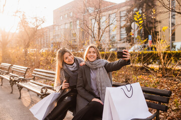 Summer sunny day, two young laughing women sitting on a park bench, rest after shopping and using their smartphones. Nearby are shopping bags. Two friends sitting in park and taking selfie.