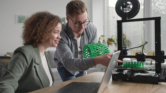 modern technologies for home, smiling man and woman print a prototype of a 3d model using a 3d printing machine sitting in a living room