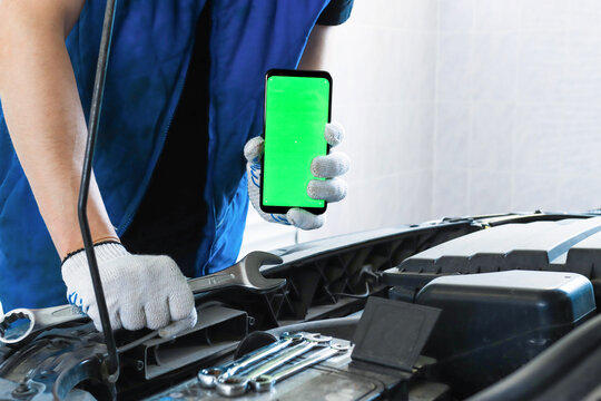 A mechanic stands near a car with an open hood in the garage and holds a key and tools and a smartphone with a chroma key. Copy space and blank. Car repair in a car service.