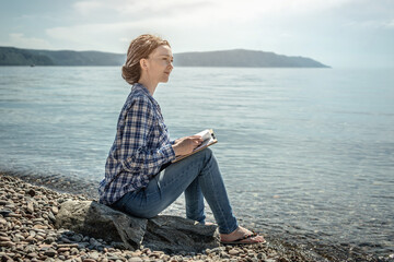 Young woman is sitting on the stony coast of a large pond, reading and enjoying the solitude and...