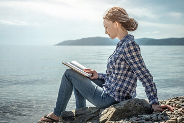 Young woman is sitting on the stony coast of a large pond, reading and enjoying the solitude and...