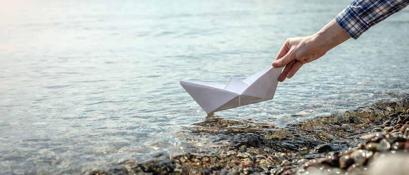 Woman's hand is launching a white paper boat into the clear water of a large lake with a stony bottom