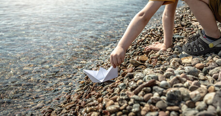 Child is launching a white paper boat into the clear water of a large lake with a stony bottom and shore
