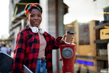 Portrait of happy african-american woman with skateboard. Young stylish woman drinking coffee outdoors.