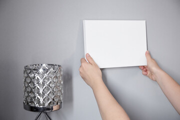 White canvas in female hands with gray wall background. Woman hanging blank picture mockup on wall