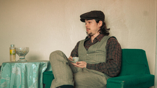 man in vintage suit is sitting on sofa and holding coffee cup. an old house. old colors. vintage background