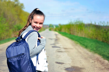 A young girl with a blue backpack turned around and smiling looks at the camera squinting at the sun against the background of a blue sky, stretching into the distance of the road plants and trees.