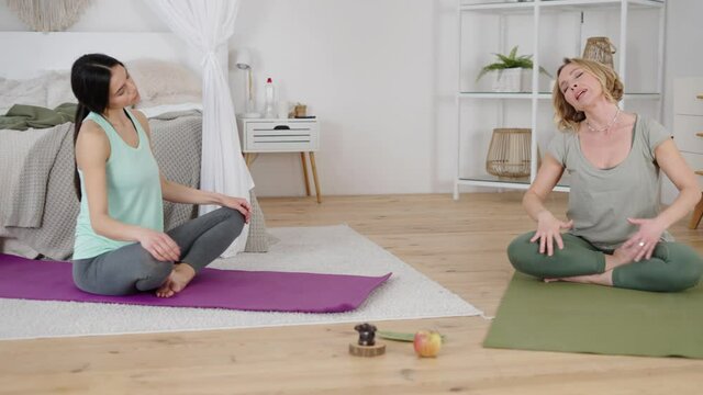 Yogs instructor explaining young woman how safely stretch neck during yoga practice at home, sitting in lotus pose on mat with closed eyes, talkig about her feelings in body, feeling calm and relaxed