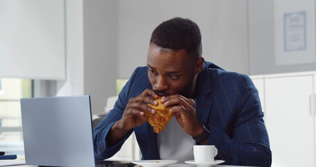 Young bearded afro businessman drinking coffee and eating croissant in modern office