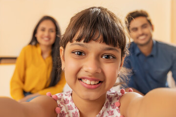 A HAPPY GIRL CHILD CLICKS A PHOTO OF HERSELF WHILE PARENTS SIT IN BACKGROUND	