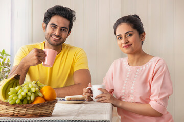 A HAPPY COUPLE CHEERFULLY LOOKING AT CAMERA WHILE SITTING WITH MORNING BREAKFAST	
