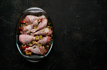Raw chicken legs and lots of spice in a transparent dish on a black background.
