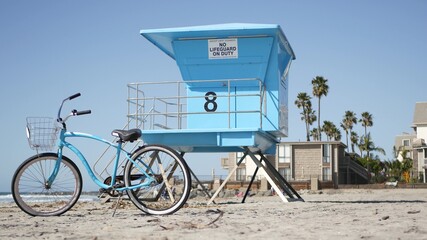 Blue bicycle, cruiser bike by ocean beach, pacific coast, Oceanside California USA. Summertime vacations, sea shore. Vintage cycle on sand near lifeguard tower or watchtower hut. Sky and water waves. - obrazy, fototapety, plakaty
