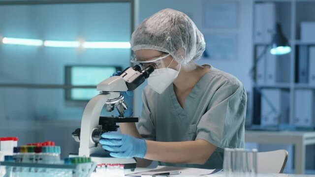 Female lab worker in protective gloves, mask, glasses and hat looking through microscope while conducting research