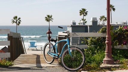 Blue bicycle, cruiser bike by ocean beach, pacific coast, Oceanside California USA. Summertime vacations sea shore. Vintage cycle by wooden stairs, stairway or staircase. Tropical palms and lifeguard - obrazy, fototapety, plakaty