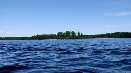 Sailing along the bright blue little waves past an island with green tree. Wild nature. Blue lake on a sunny day. Blue sea and sky with clouds. Summer and travel vacation concept.