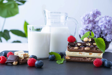 broken bars of black, white and milk chocolate with nuts blueberries and raspberries, a jug of milk
