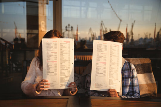 Young funny happy loving couple sitting at sea port open-air cafe and fooling closing their faces with menus. Beginning of love story. Relationship love, lifestyle concept. Couple in love on a date
