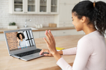 Smart mixed-race teenage girl student or freelancer in glasses sitting on the couch with laptop on lap, studying from home and waving at the screen, watching an online webinar, saying hi, taking notes