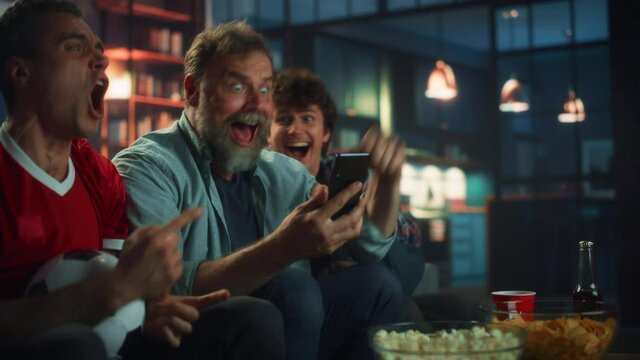 Night at Home: Three Soccer Fans Sitting on a Couch Watch Game on TV, Use Smartphone App to Online Bet, Celebrate Victory when Sports Team Wins. Friends Cheer Eat Snacks, Watch Football Play.