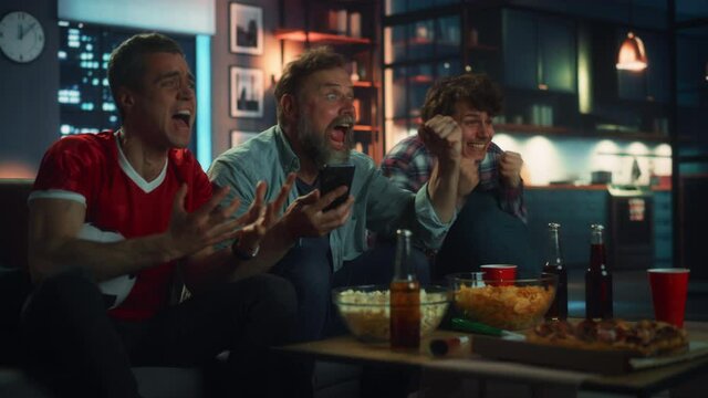 Night at Home: Three Soccer Fans Sitting on a Couch Watch Game on TV, Use Smartphone App to Online Bet, Celebrate Victory when Sports Team Wins. Friends Cheer Eat Snacks, Watch Football Play. Wide