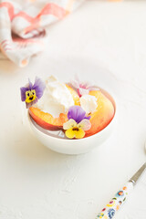 Peaches served in white plate with mascarpone, pink flower, violets and herbs on the white background. Summer dessert.