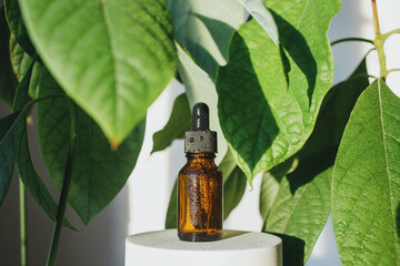 A brown glass bottle of serum, essential oil or other cosmetic product, among large avocado leaves....