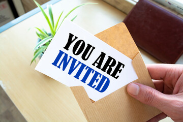 Male hand holding craft envelope with text YOU ARE INVITED on blue background