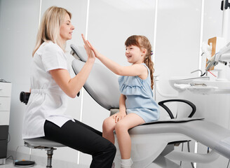 Cute little girl and female dentist giving high five after dental procedure. Smiling child sitting...