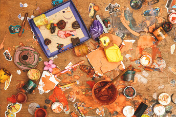 Jars of colorful paints brushes plasticine pieces and small toys on messy wooden table in light children room upper view