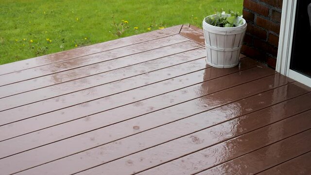 A brown composite outdoor floor with wood pattern. Heavy rain causes raindrops to fall on the floor. In the background is a green lawn with yellow dandelions. A white large pot with green leaves.