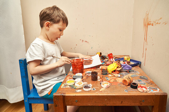 Surprised little boy draws picture with brush at messy table