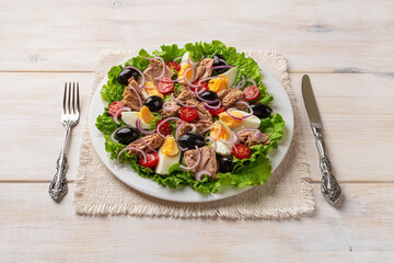 Tasty tuna salad on a white plate, knife and fork over white wooden table. Ready to eat mediterranean tuna salad with boiled eggs, fresh lettuce and tomatoes. Omega-3 dietary healthy eating.