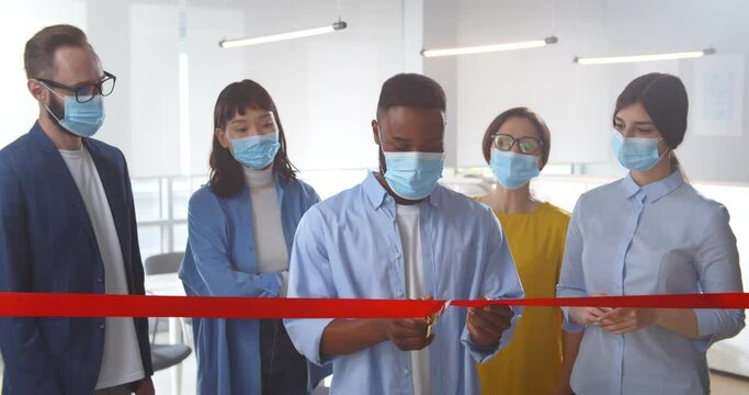 Happy business team in safety mask cutting red ribbon in office celebrating reopening