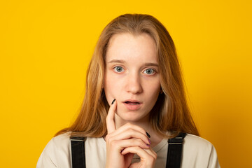 Pensive teen girl being deep in thoughts, raises eyebrows, curves lips, holds chin.