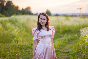 Beautiful portret of happy woman with dark hair standing in the wildflowers field holding the bottom of the long pink dress on a warm summer evening. Joyfull summer walking. 