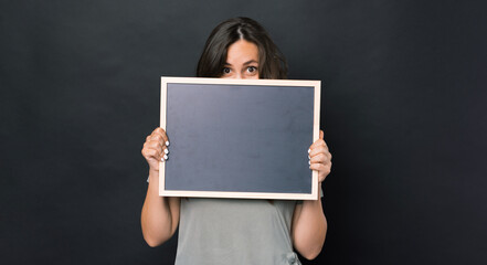 Photo of amazed woman holding black empty chalk board over dark background and covering face