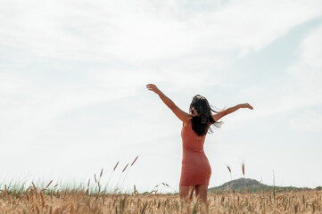 Side view of slender young lady rising her arms up to Sun, having walk in wheat field, wearing a...
