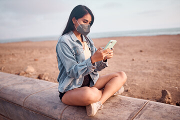 Mujer Joven latina revisa su celular durante un bello atardecer.. Joven feliz  con mascarilla en la playa
