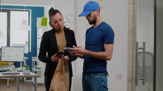 Delivery man bringing takeaway food meal order to businesswoman in startup company office during takeout lunchtime. Entrepreneur receiving lunch paying with smartphone using POS contactless service