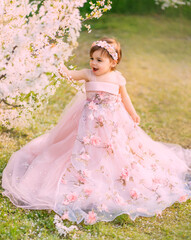 Obraz na płótnie Canvas Young happy little girl fairy princess. Happy child face is smiling. Luxurious fluffy long dress for children, pink outfit. White flowers background trees spring nature. Fashion model, one year old.