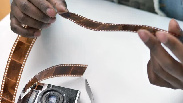 Smiling African American guy looks at small outdated brown photographic film near historical camera on white studio table close view