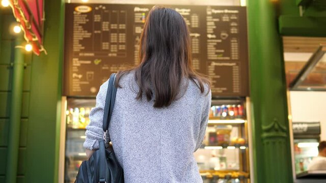 Back view of the girl, the buyer looks at the menu on the wall in the cafe, chooses a drink. Selects a dish and drinks to order.