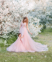 Young happy family, mom and daughter in beautiful evening festive long powdery pink color dresses. Woman holds little girl in hand, looks with love, smile on face. Wedding lush outfits. Nature spring.