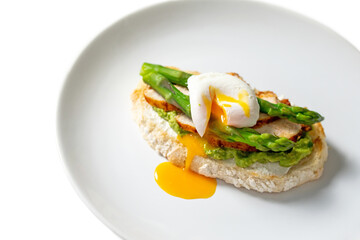 Instruction, step 7. Tasty morning toast with cream cheese, mashed avocado, roasted chicken slices, asparagus and poached egg with bright yolk on white plate isolated. Healthy breakfast recipe. 