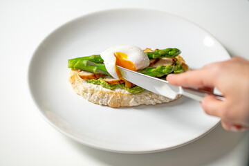 Hand holding knife and cutting poached egg on delicious morning toast with cream cheese, mashed avocado, roasted chicken slices, asparagus on white plate. Healthy nutritious breakfast recipe. 