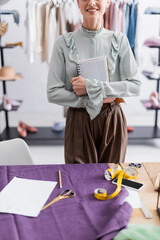 Cropped view of smiling seamstress holding notebook near cloth, tape measure and cellphone